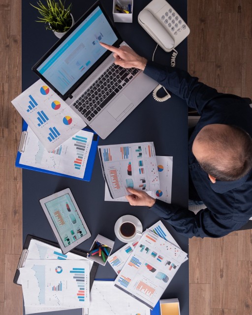 Man in white winter jacket and goggles crouching beside a Dalmatian dog wearing sunglasses and fluffy earmuffs in a snowy mountain landscape.